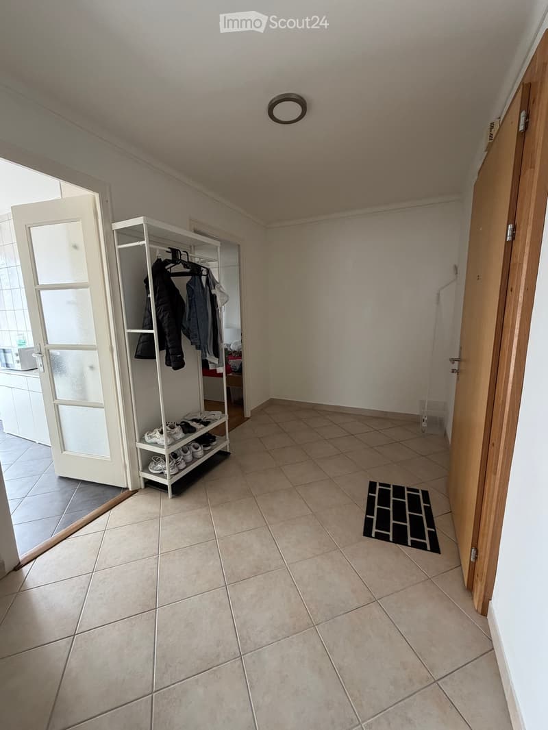 Empty hallway with tiled floor, white walls, and a small shoe rack holding shoes and jackets. A black doormat is placed near the door on the right.