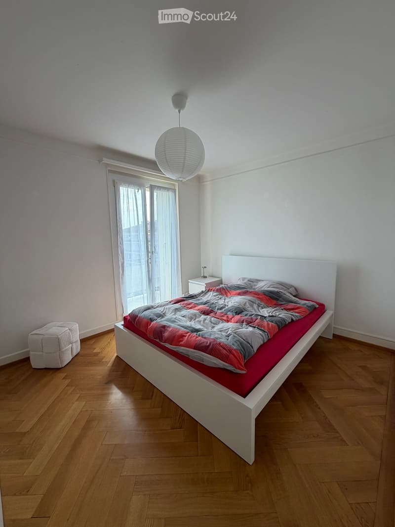Modern bedroom with a white platform bed, red and grey duvet, white headboard, wooden floor, and a hanging paper lantern light fixture.