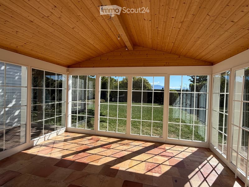 Bright sunroom with wooden ceiling, large glass windows, and terracotta tile flooring, offering a view of a green lawn and distant mountains.