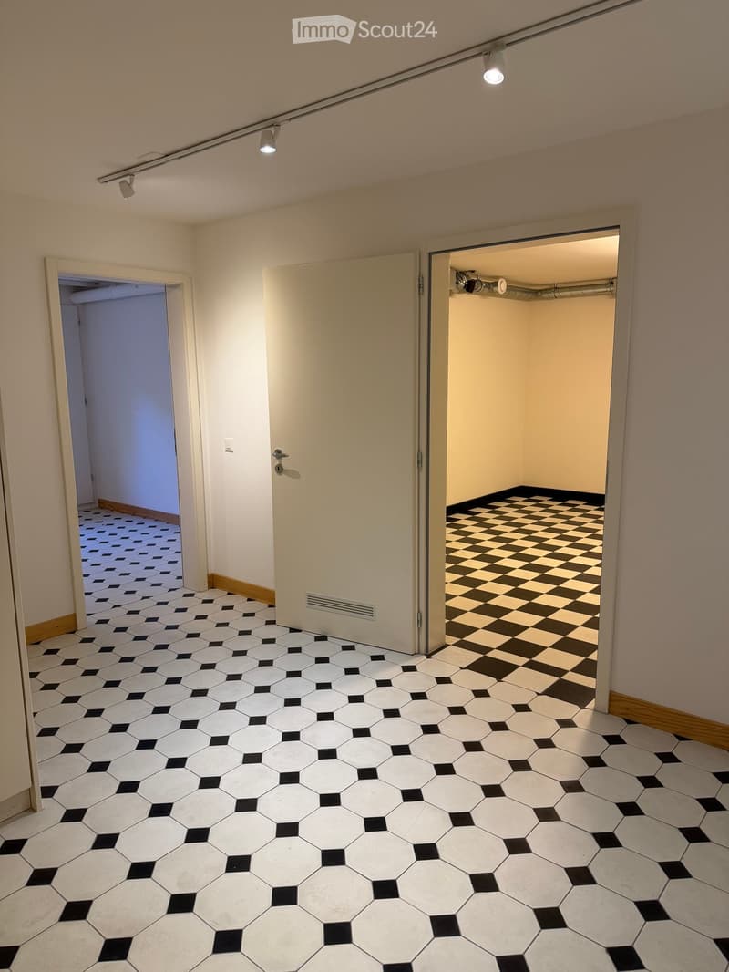 Empty hallway with hexagonal white and black tiled floor, beige walls, and three doors leading to different rooms.