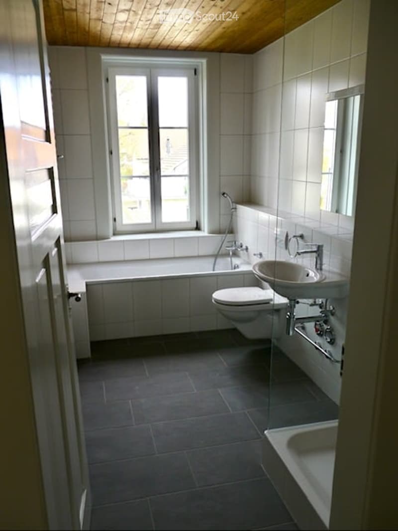 Modern bathroom with white tiles, a bathtub, toilet, sink, and a large window providing natural light.