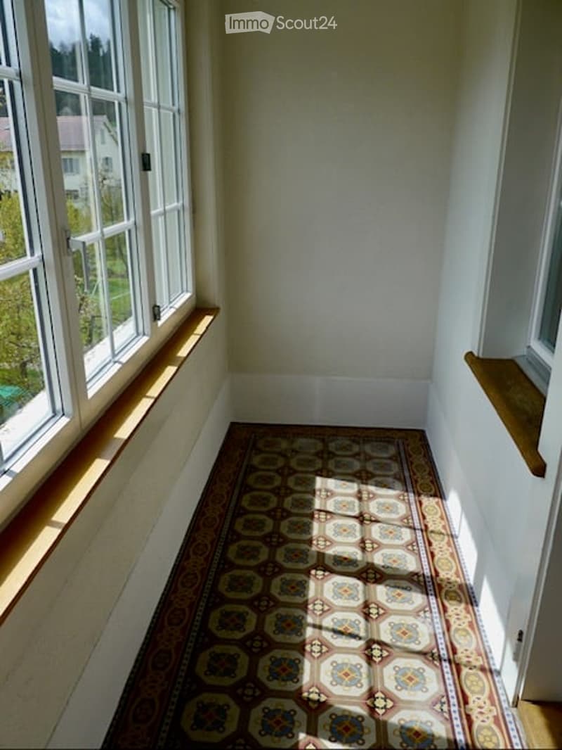 Empty room with large windows, wooden floor, and patterned rug. No furniture or equipment visible.