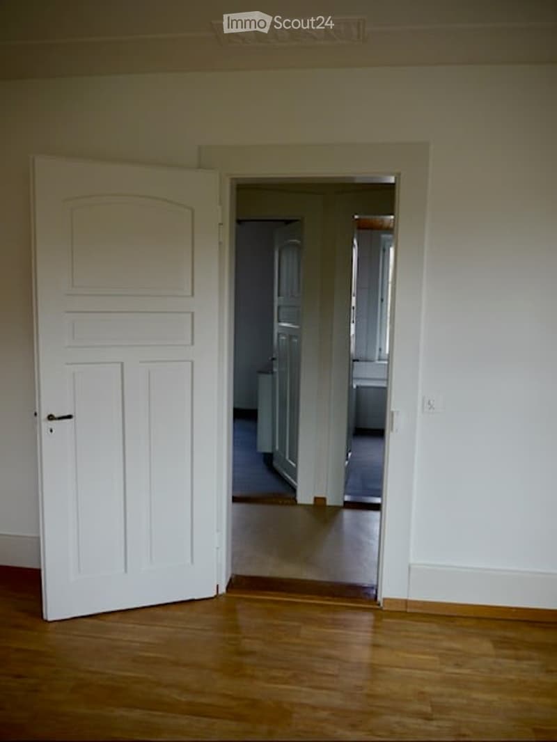 Empty hallway with wooden flooring, white walls, and an open door leading to another room.
