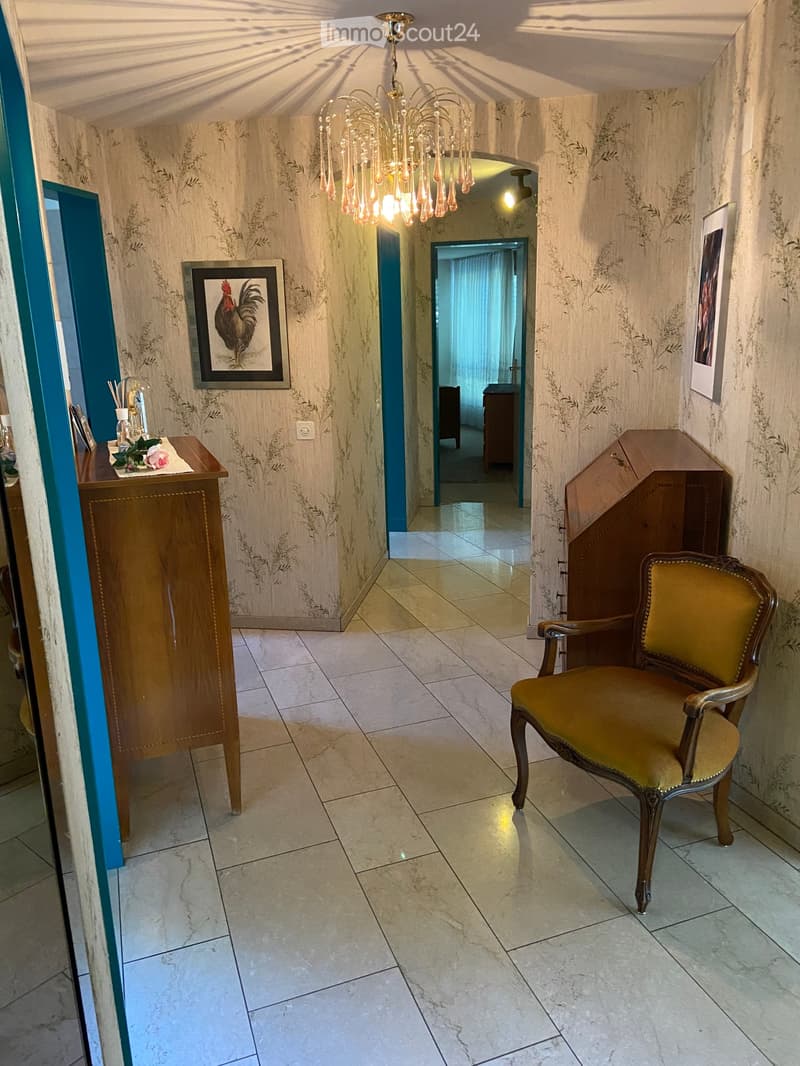 Elegant hallway with patterned wallpaper, a chandelier, a small wooden table with a flower arrangement, a framed picture, and a yellow upholstered chair near a wooden cabinet.