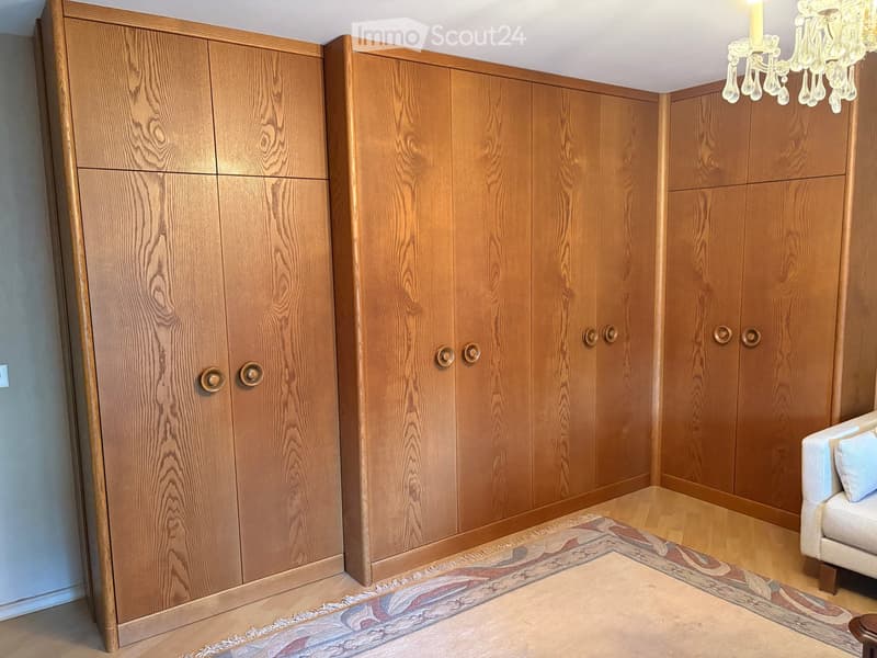 A well-lit room with wooden wardrobes, a light-colored sofa, a patterned rug, and a chandelier hanging from the ceiling.