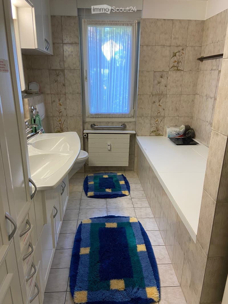 Bathroom with a sink, toilet, bathtub, and two rugs on the floor. The walls are tiled with a floral design, and there is a window with a curtain above the bathtub.