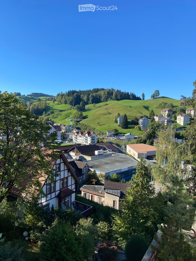 A panoramic view of a town nestled in green hills, with houses, trees, and a clear blue sky.
