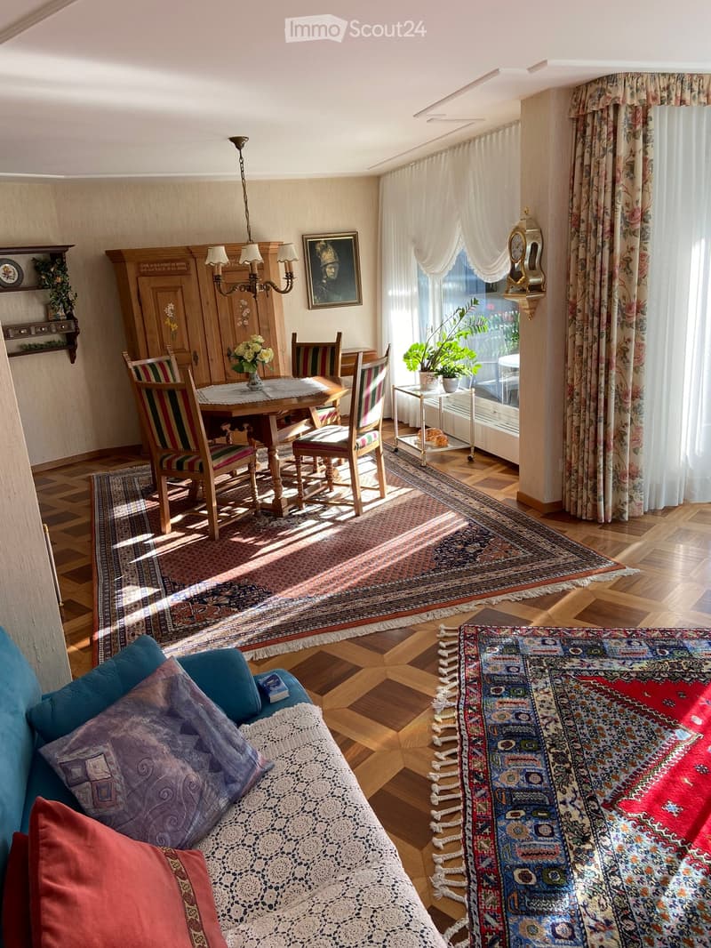 A well-lit living and dining area with wooden floors, a large patterned rug, a sofa with colorful cushions, a dining table with chairs, and a chandelier hanging from the ceiling.