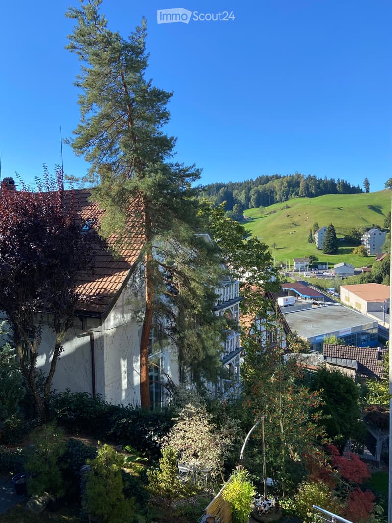 View from a balcony or terrace showing a garden area with various plants and trees, a white building with a tiled roof, and a scenic countryside with green hills and scattered houses in the background.