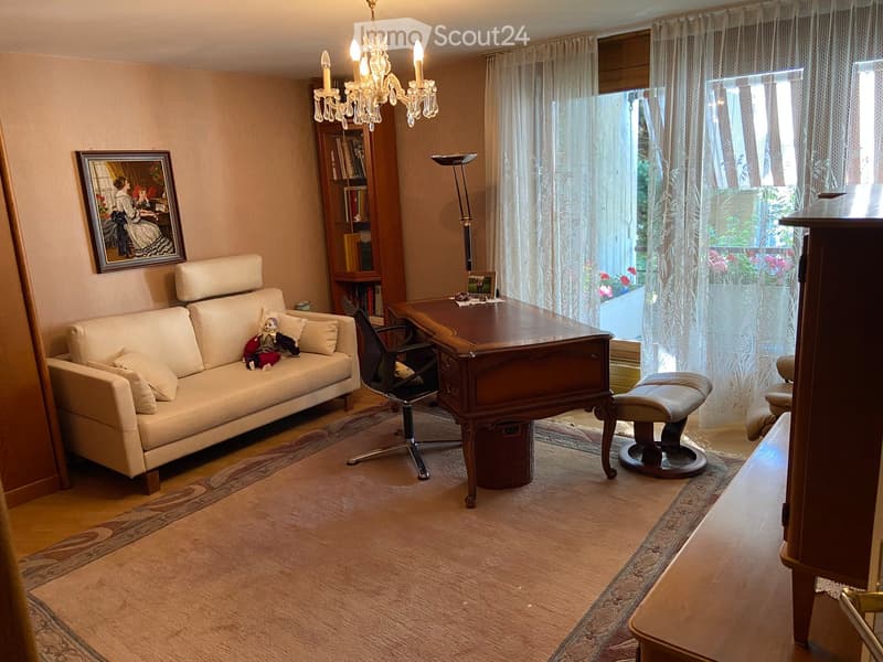 A well-furnished living room with a cream-colored sofa, a wooden desk, a chair, a small stool, a bookcase, a chandelier, and a picture frame on the wall. The room has large windows with sheer curtains and a patterned rug on the floor.