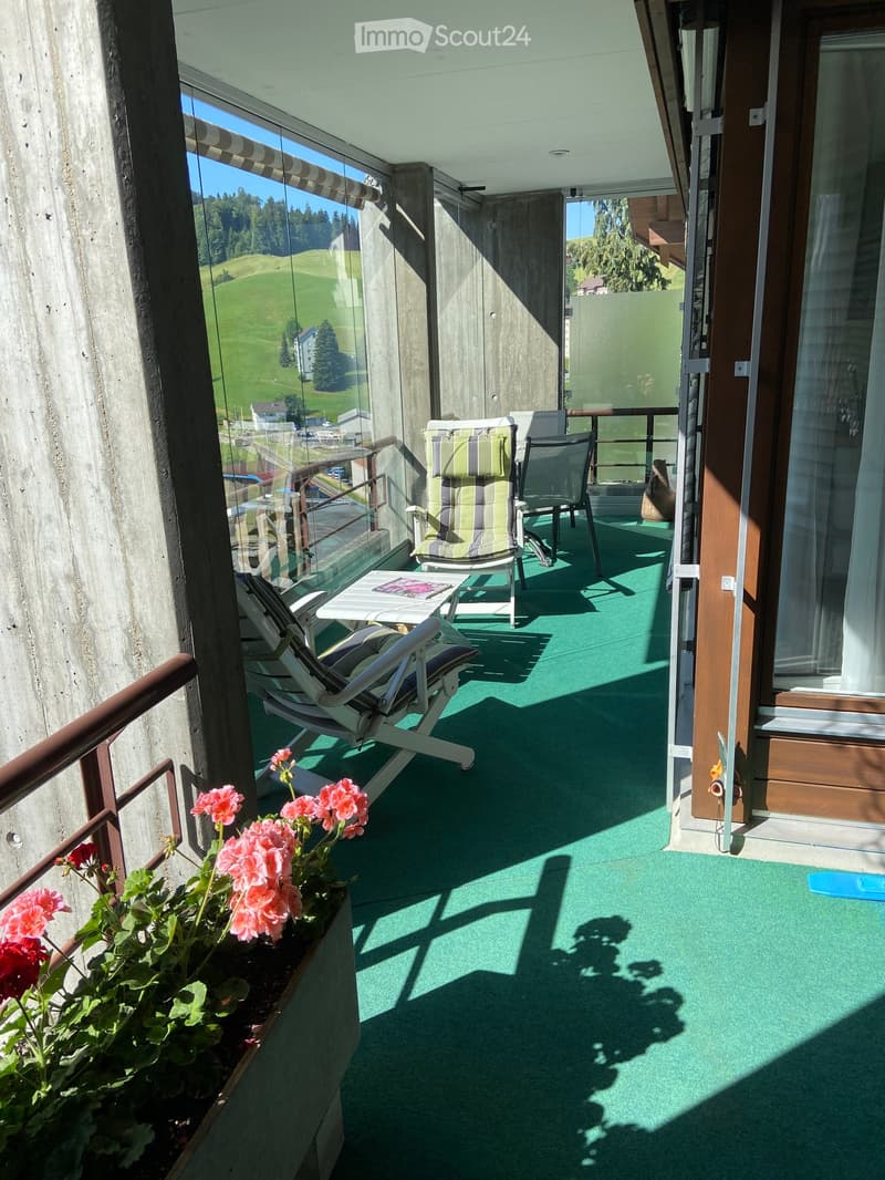 Balcony with green carpet, chairs, small table, and flowers, overlooking a green landscape.