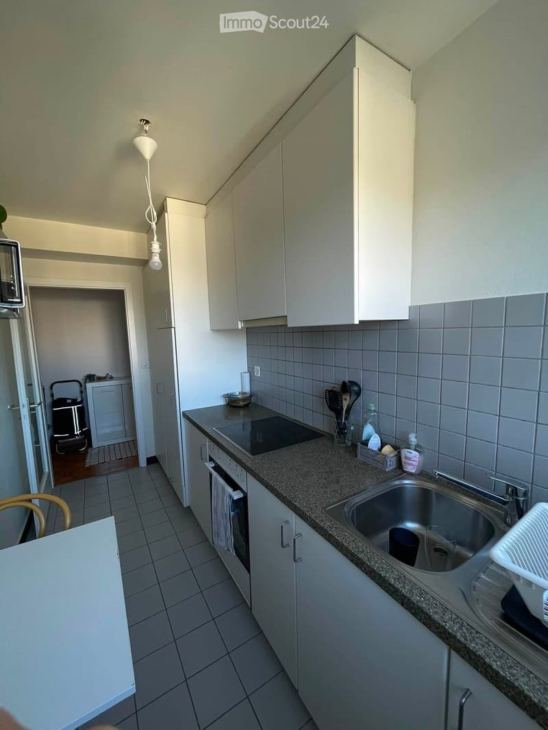 Modern kitchen with white cabinets, granite countertop, stainless steel sink, and built-in oven. Visible items include a dish rack, kitchen utensils, and a refrigerator in the background.
