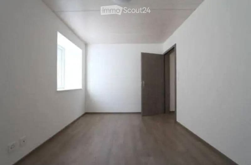 Empty room with white walls, wooden floor, and a single window on one side. A closed door is visible at the far end.