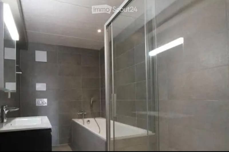 Modern bathroom with a bathtub, sink, and glass shower enclosure. The walls are tiled in a neutral color, and the lighting is bright.