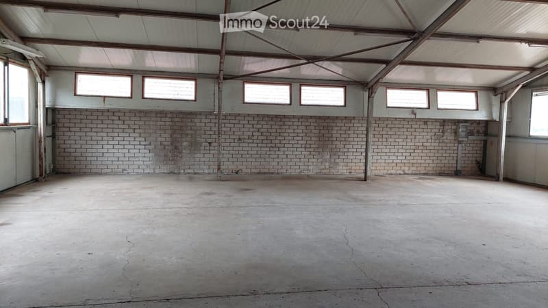 Empty warehouse or garage space with exposed wooden beams, brick walls, and large windows, showing a concrete floor and no furniture or equipment.