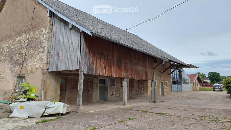 A rustic barn with a wooden exterior and stone walls, featuring a large open doorway and a covered entrance area. The surrounding area includes a paved ground and some scattered equipment.
