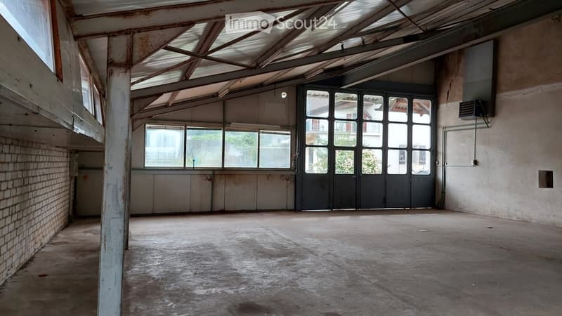 Empty industrial-style room with exposed metal ceiling beams, large windows, and a central metal door. Bare concrete floor and brick walls.