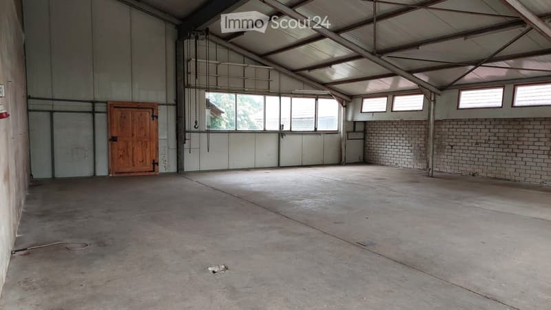 Empty warehouse or garage with concrete floor, metal walls, wooden door, and large windows allowing natural light.