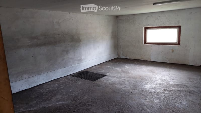 Empty room with bare concrete walls and floor, featuring a single window and a metal floor drain.