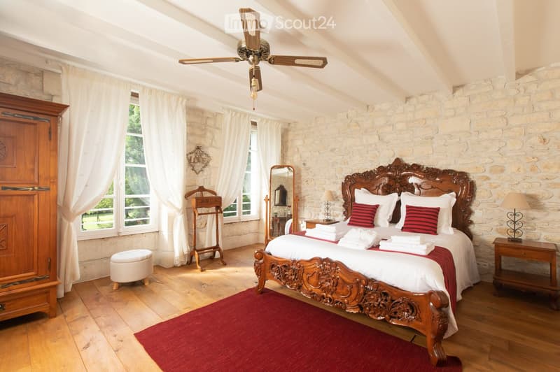 Cozy bedroom with wooden bed, nightstands, lamps, and a ceiling fan. Features stone walls, wooden flooring, and large windows with white curtains.