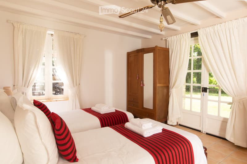 Bright bedroom with two single beds, red and black striped bedspreads, white pillows, a wooden wardrobe, and large windows with white curtains.