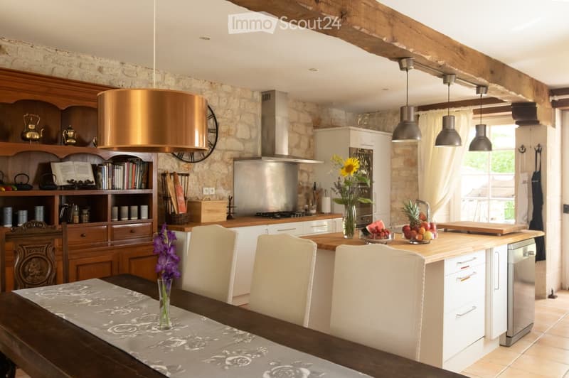 A rustic kitchen with exposed stone walls, wooden beams, and a central island. The space features a dining table with chairs, a wooden cabinet with glass doors, and various kitchen appliances including a dishwasher. There are decorative items such as vase