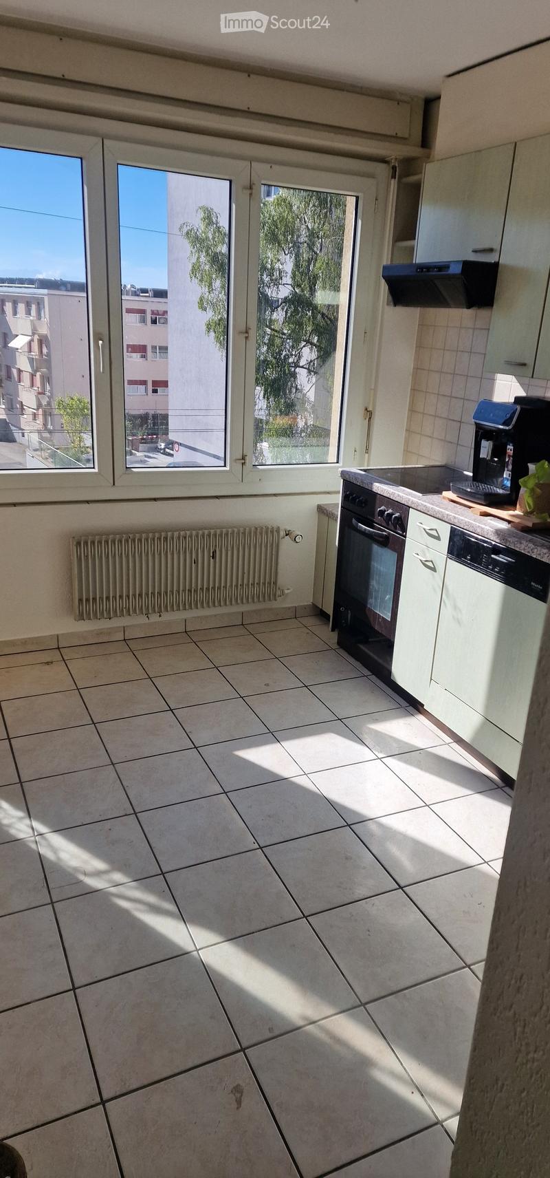 Bright kitchen with tiled floor, white cabinets, oven, dishwasher, coffee maker, and radiator. Large windows provide natural light.