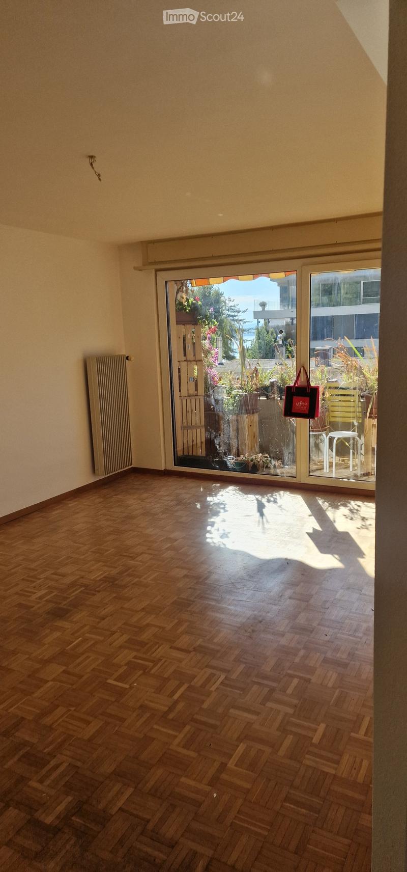 Empty room with wooden herringbone flooring, large sliding glass doors leading to a balcony area with outdoor furniture and plants visible.