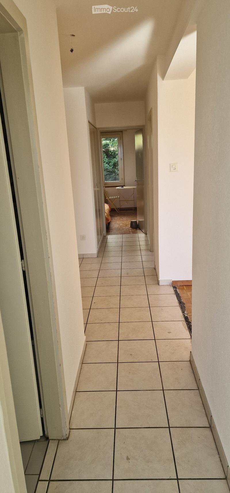 A long, narrow hallway with tiled flooring and white walls, leading to a room with a small table and chair visible at the end.