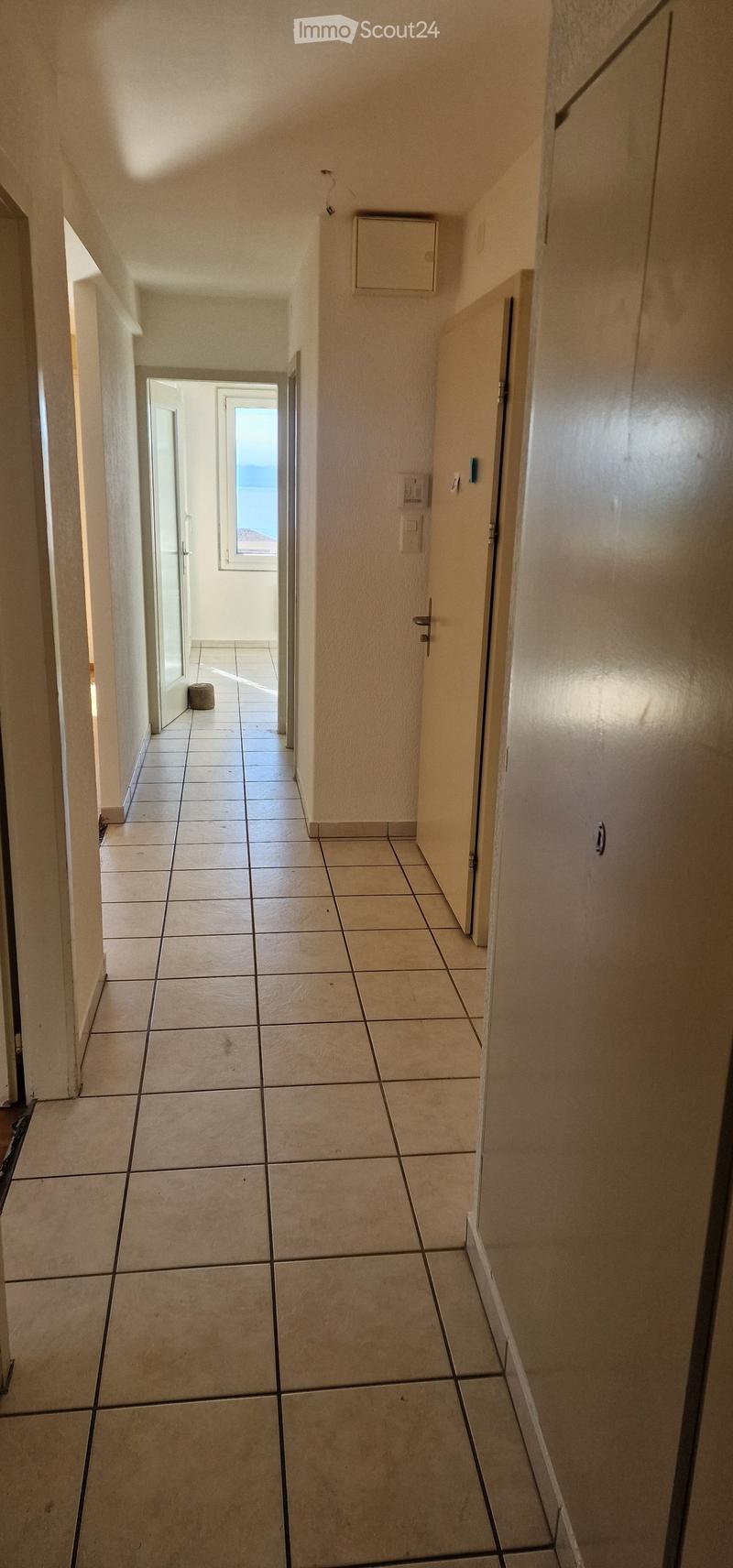 Empty hallway with tiled floor, white walls, and a window at the end showing a view of the sea.