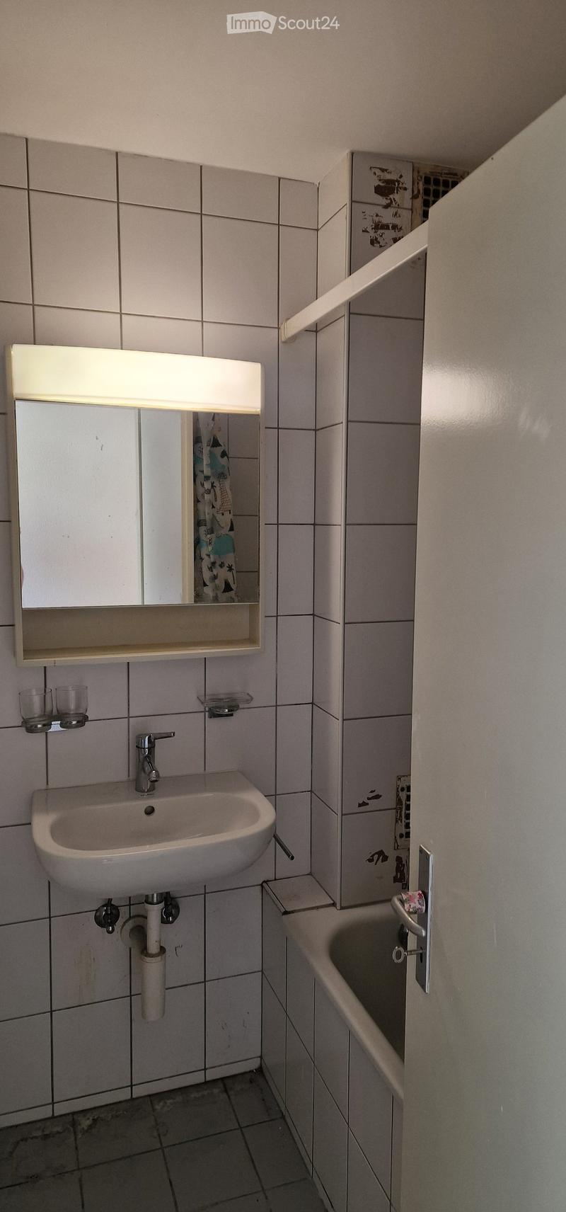 Small bathroom with a white tiled wall, a sink with a faucet, a mirror with built-in lighting, two cups on a shelf, and a bathtub with a handle.
