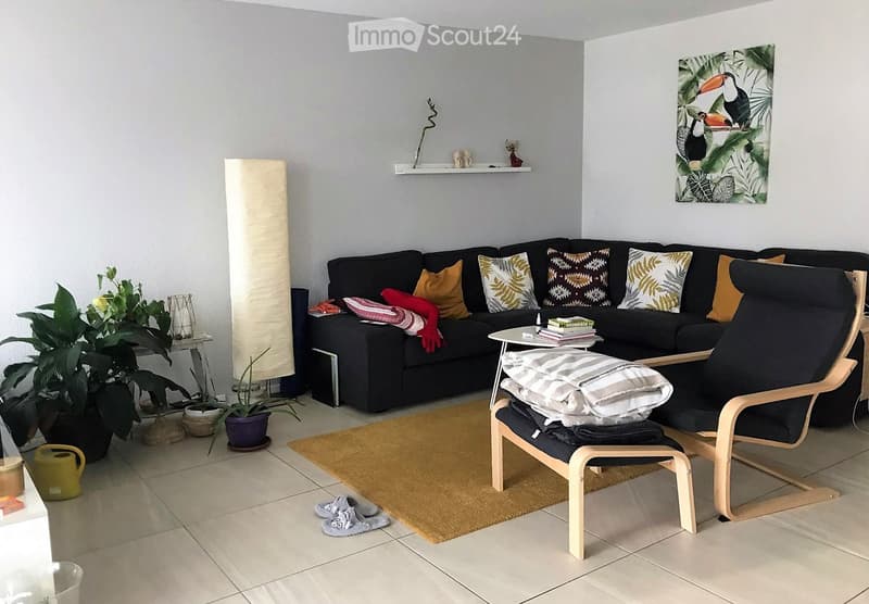 Modern living room with a black sectional sofa, colorful throw pillows, a wooden chair with folded blankets, a small side table with books, a potted plant, and a large floor lamp.