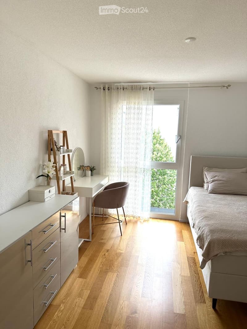 Modern bedroom with a bed, a wooden desk, a chair, a mirror, and a flower vase on a white shelf.