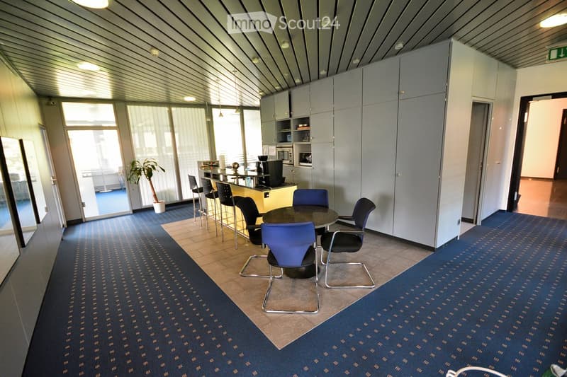 Modern office space with a round table surrounded by chairs, a bar area with stools, cabinets, and a microwave. Blue carpeting and large windows provide natural light.