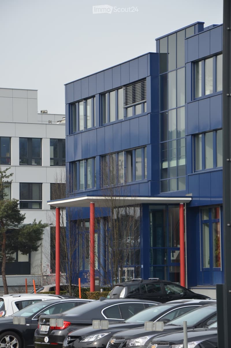 Modern two-story building with blue facade and large windows, surrounded by parked cars and trees.