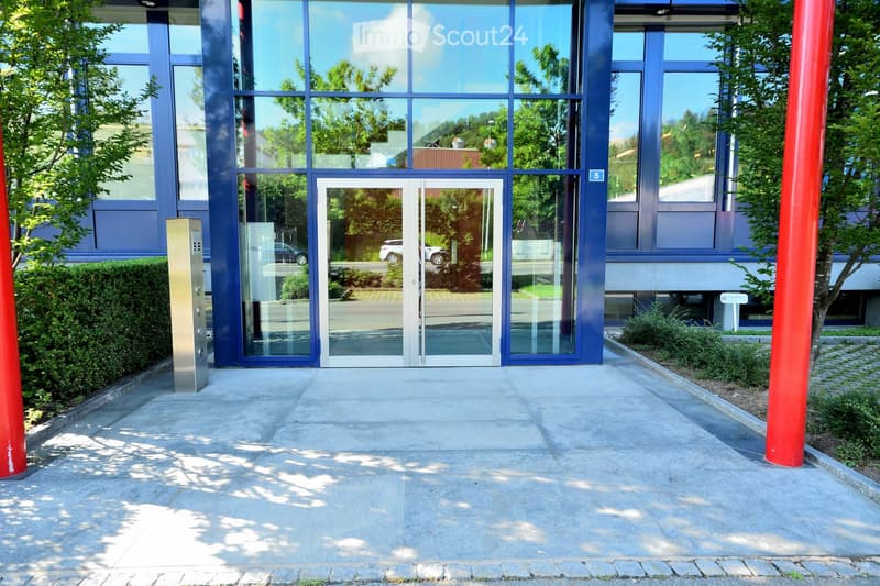 Modern building entrance with glass doors and blue framing, surrounded by greenery and a paved walkway.