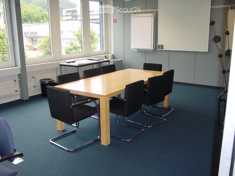 Meeting room with a large wooden table, black chairs, and a whiteboard on the wall. Large windows provide natural light, and the room has a blue carpeted floor.