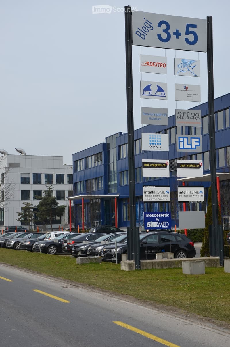 Modern multi-story building with blue facade and numerous company logos displayed on a signpost. Several cars are parked in the lot in front of the building.