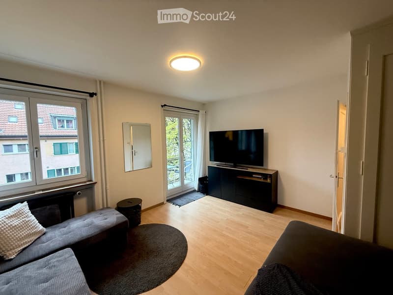 Bright living room with wooden flooring, large windows, balcony access, sofa, ottoman, coffee table, TV, and cabinet.