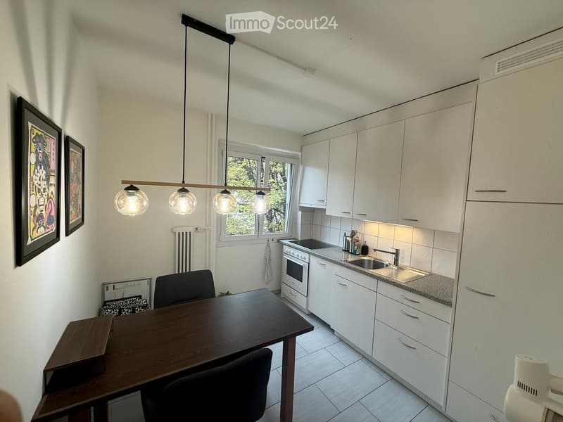 Modern kitchen with white cabinets, granite countertop, built-in oven, sink, and dining area with a wooden table and chairs. Features pendant lighting and large windows with a view of greenery.