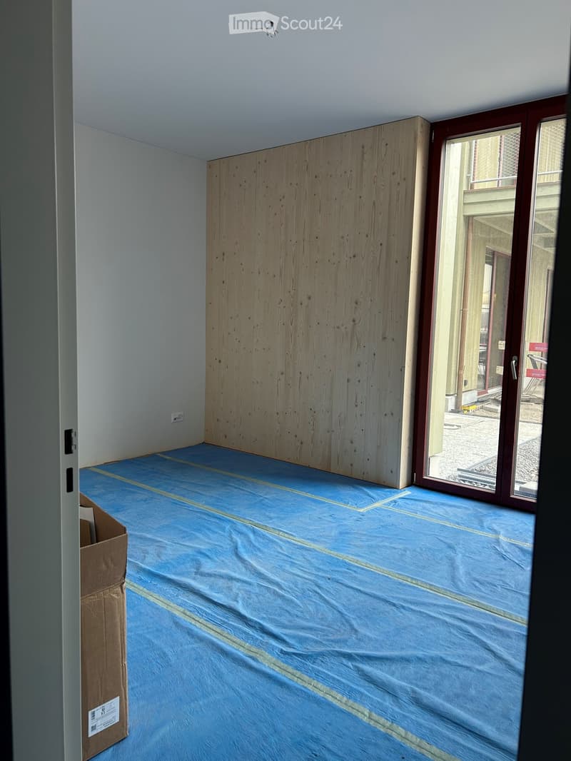 Empty room with blue protective flooring and a wooden panel wall. A cardboard box is visible in the corner, and a large window with a view to an outdoor area is present.