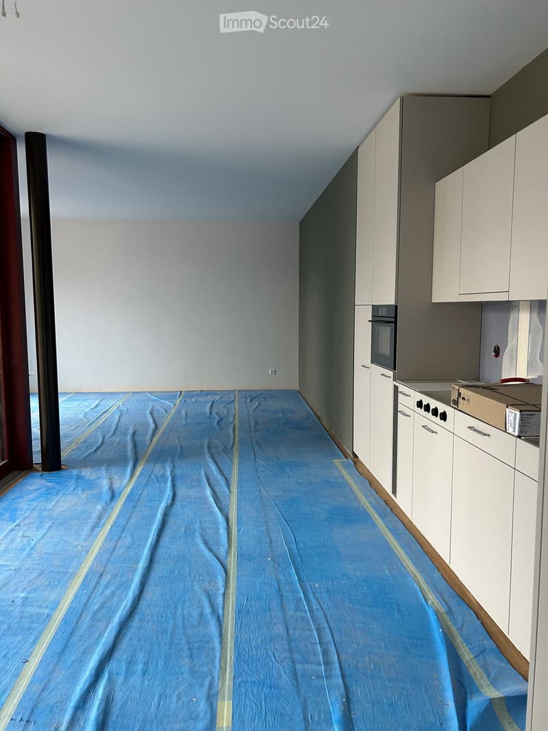 Empty room with white walls, a blue protective floor covering, and white kitchen cabinets along one wall.
