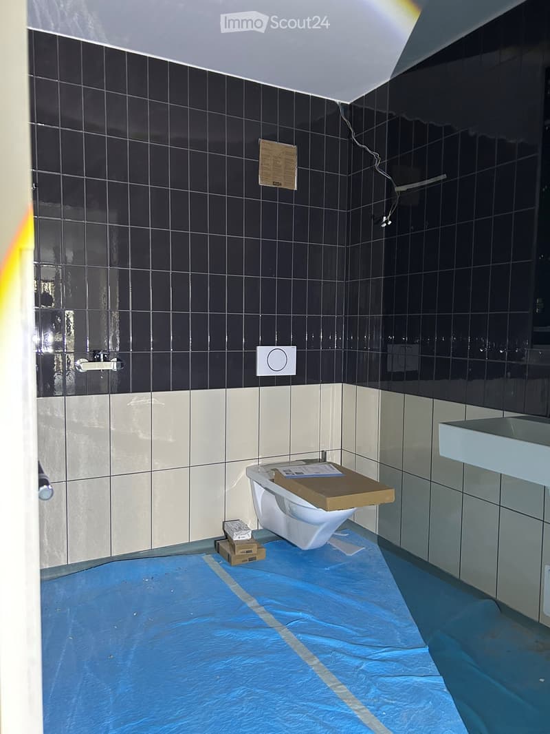 Bathroom with black tiled walls, white lower tiles, a toilet with a cardboard box on top, a sink, and a blue protective floor covering.