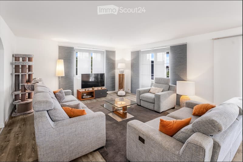 Modern living room with grey sofas, armchairs, a glass coffee table, TV stand, floor lamp, table lamp, and a decorative shelf.