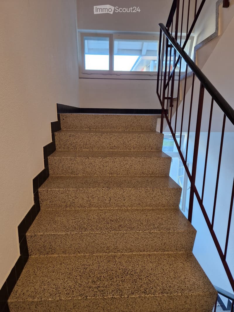 Staircase with beige tiled steps and a black metal railing, leading up to a window at the top.