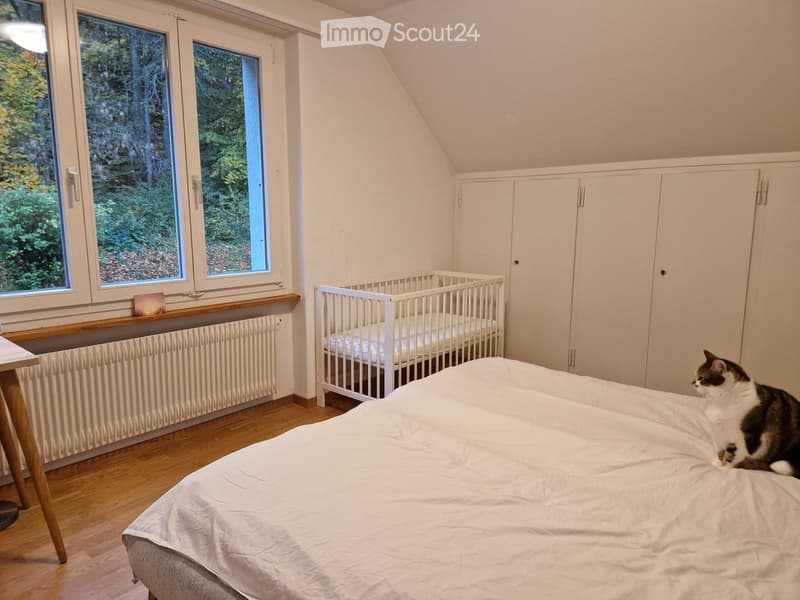 Cozy bedroom with a large bed, a small crib, built-in white cabinets, wooden flooring, and large windows overlooking a green landscape.