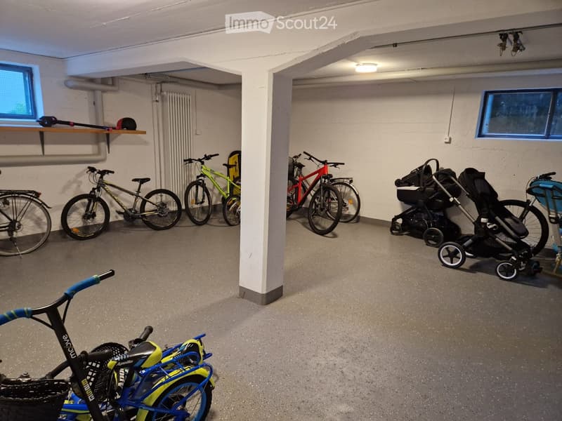 Garage with multiple bicycles and a baby stroller, concrete floor, white walls, small windows, and a shelf with items on the left side.
