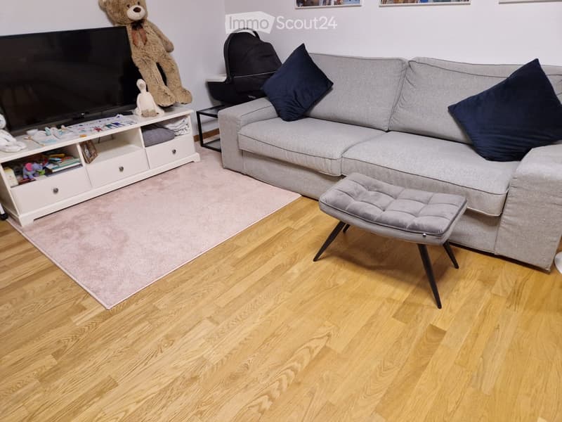 Modern living room with a grey sectional sofa, dark blue throw pillows, a grey ottoman, a TV stand with a flat-screen TV, and a small table with toys and a backpack.