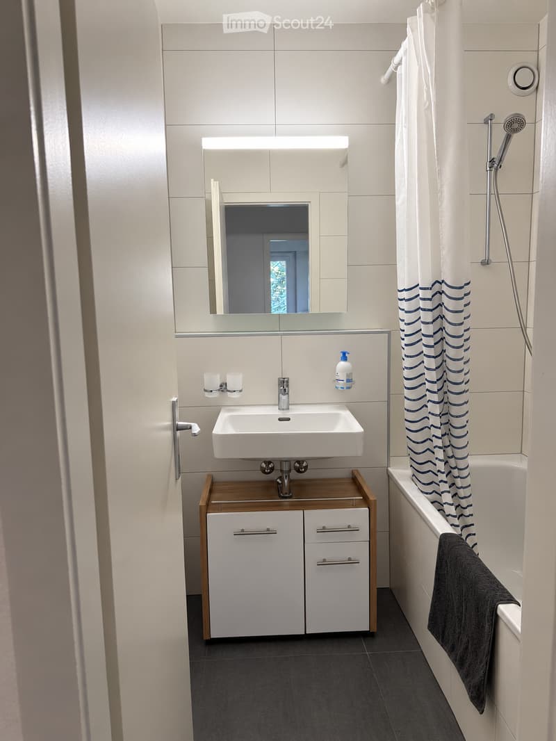 Modern bathroom with white tiles, a white sink with a wooden cabinet, a bathtub with a blue-striped shower curtain, and a towel draped over the edge.