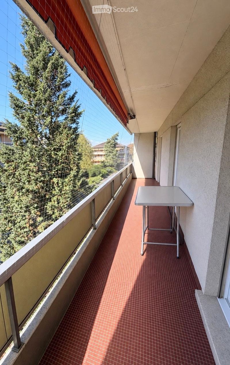 Balcony with red tiled floor, white table, large windows with blinds, and a view of trees and buildings.